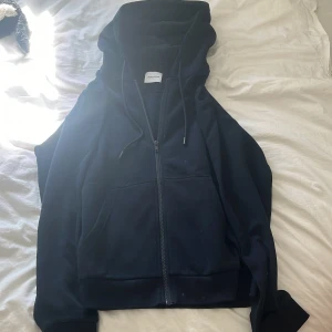 Mörkblå hoodie med dragkedja - Säljer en mörkblå hoodie från Jack & Jones med hel dragkedja framtill, huva med snörning och två stora fickor. Perfekt för chill dagar eller när du vill ha en enkel och stilren look. Materialet är mjukt och skönt, troligtvis bomullsmix.