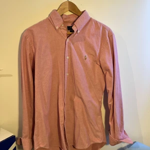 Rosa skjorta från Polo Ralph Lauren - Snygg rosa skjorta från Polo Ralph Lauren i slim fit-modell. Skjortan har klassisk krage, knäppning framtill och broderad logga på bröstet. Tillverkad i bomull och har långa ärmar med manschettknappar. Perfekt för en clean och stilren look.