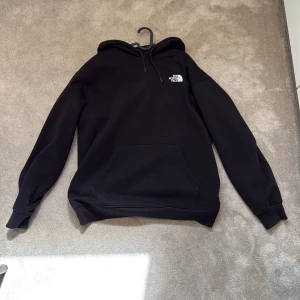 Svart hoodie från The North Face - Svart hoodie från The North Face med vit logga på bröstet. Klassisk känguruficka framtill, huva med snörning och ribbade muddar. Perfekt för chill dagar eller när du vill ha en enkel och stilren look. Använd 1-3 gånger