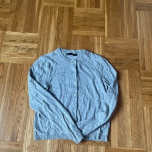 Ljusgrå kofta med knappar - En stilren ljusgrå kofta med rund hals och knäppning framtill. Koftan har långa ärmar och ribbade muddar vid ärmslut och nederkant. Perfekt att slänga över en t-shirt eller klänning för en chill look.