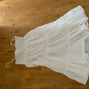 Vit långklänning med smock och volang - Superfin vit långklänning med smock upptill och tunna axelband. Klänningen har volangig kjol och är gjord i ett lätt, luftigt tyg som passar perfekt till varma dagar. Enkel och stilren design som känns både romantisk och trendig. Etikett saknas men sitter som en S, finns en liten fläck (se på bilden) men den syns knappt