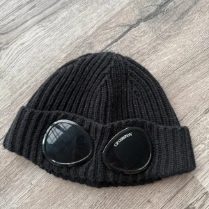 Mössa C.P. Company - Svart ribbstickad mössa från C.P. Company med ikoniska svarta glasögonlinser framtill på uppviket. Mössan är tillverkad i mjukt stickat material och har en modern streetwear-look som sticker ut. Skick 10/10. Skicka pm vid minsta fundering