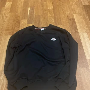 Svart sweatshirt från Moncler - Svart sweatshirt från Moncler i storlek S. Tröjan har rund halsringning, ribbade muddar och ett broderat Moncler-logo på bröstet. Enkel och stilren design som passar perfekt till jeans eller joggers. Materialet känns mjukt och bekvämt mot huden.