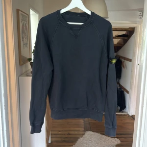 Svart Stone Island sweatshirt - Svart sweatshirt från Stone Island med klassisk rund hals och ribbade muddar. Tröjan har den ikoniska loggan på vänster ärm och är tillverkad i mjuk bomull. Perfekt för dig som gillar streetwear och vill ha en clean look.