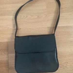 Svart axelväska i skinnlook - En stilren svart axelväska med enkel design och mjuka former. Väskan har ett lock framtill och en praktisk dragkedjeficka på baksidan. Perfekt storlek för det viktigaste och lätt att bära över axeln. Materialet har en snygg skinnlook.