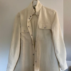 Beige manchester overshirt - Snygg beige overshirt i manchester med raka linjer och tryckknappar framtill. Jackan har två bröstfickor med lock och lång ärm. Perfekt för lager-på-lager och ger en chill vibe till din outfit.