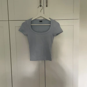 Ljusblå ribbad topp från Gina Tricot - Supersöt tajt ljusblå ribbad topp från Gina Tricot i storlek XS. Modellen är croppad med korta ärmar och har en bred, rundad urringning som ger en snygg siluett. Perfekt att matcha med jeans eller kjol för en fräsch look.