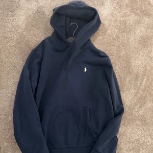 Marinblå hoodie från Polo Ralph Lauren - Snygg marinblå hoodie från Polo Ralph Lauren med klassisk broderad logga på bröstet. Tröjan har huva med snörning och en stor känguruficka framtill. Perfekt för dig som gillar stilren och avslappnad look.