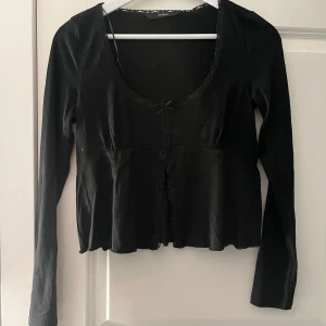 Svart långärmad blus från Vero Moda - Supersöt svart blus från Vero Moda med lång ärm, söt liten rosett framtill och spetsdetaljer längs urringningen och knäppningen. Modellen är lätt utsvängd nertill och har en feminin, figurnära passform. Perfekt att styla med jeans eller kjol.