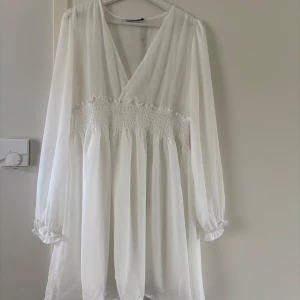 Vit omlottklänning med smock i midjan - Superfin vit kortklänning med omlottliv, smockad midja och långa ärmar med volangkant. Klänningen har en luftig känsla och v-ringning, perfekt för en fräsch och stilren look. Materialet är lätt och flowy, vilket gör den extra bekväm. Oanvänd med prislapp kvar strl L