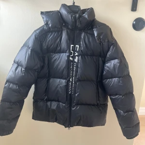 Svart pufferjacka från Emporio Armani - Säljer en svart pufferjacka från Emporio Armani med hög krage, huva och tydlig logga på ärmen. Jackan har dragkedja framtill och breda, quiltade sektioner. Perfekt för kalla dagar och riktigt snygg streetstil. Har ett litet hål vid märket vid sidan. 