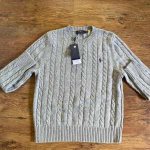 Grå kabelstickad tröja Polo Ralph Lauren - Kabelstickad grå tröja från Polo Ralph Lauren med klassisk rund halsringning och broderad logga på bröstet. Tröjan ligger på storlek S.
