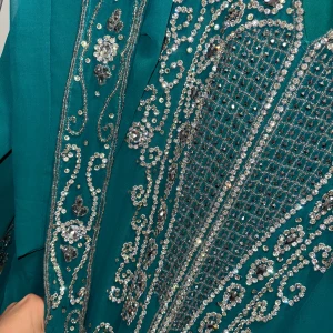 Marockans festklänning - En turkos långklänning med v-ringning och långa ärmar, täckt av glittriga paljetter och stenar i silvermönster. Klänningen har ett unikt och festligt utseende med detaljer längs ärmar och hela framsidan. Perfekt för dig som vill sticka ut och glänsa.