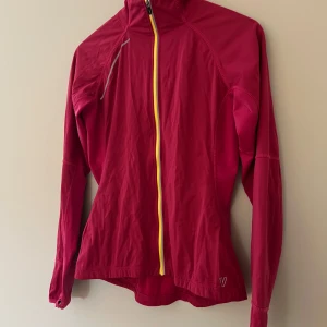 Rosa vindjacka från deVal Woman S - Sportig rosa vindjacka från deVal Woman i storlek S. Jackan har en gul dragkedja framtill, hög krage och lång ärm. Perfekt för utomhusaktiviteter och träning. Snygg passform och lätt material som andas.