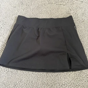 Svart sportig kortkjol från Stay In Place - Svart sportkjol från Stay In Place med inbyggda shorts under och liten slits framtill. Perfekt för dig som vill ha en bekväm och snygg look till träning eller tennis. Kjolen är i stretchigt syntetmaterial och har en bred midjeresår för extra komfort.