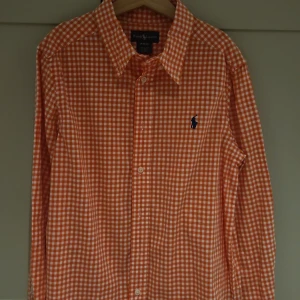 Rutig orange skjorta från Ralph Lauren - Snygg orange och vit rutig skjorta från Ralph Lauren med klassisk krage och långa ärmar. Skjortan har knäppning framtill och ikonisk blå logga broderad på bröstet. Perfekt för dig som vill sticka ut med färg och stil. Tillverkad i bomull.