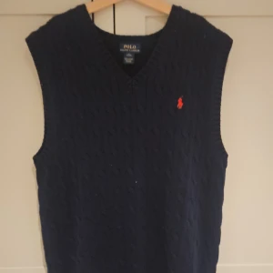 Mörkblå stickad väst Polo Ralph Lauren - Kabelstickad mörkblå väst från Polo Ralph Lauren med klassisk v-ringning och röd broderad logga på bröstet. Perfekt att slänga över en skjorta för en preppy look. Mjuk och skön känsla tack vare det stickade materialet.