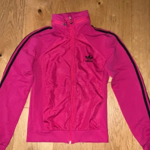 Rosa Adidas vindjacka med svarta detaljer - Säljer en snygg rosa Adidas vindjacka med svarta ränder längs ärmarna och broderad logga på bröstet. Jackan har hel dragkedja, hög krage och diskret mönster framtill. Perfekt för dig som gillar sportig stil och vill sticka ut. Retro tröja!😃