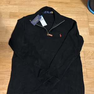 Svart half zip tröja Polo Ralph Lauren - Svart långärmad tröja från Polo Ralph Lauren med röd broderad logga på bröstet. Tröjan har half zip-dragkedja och är tillverkad i mjuk bomull. Perfekt för en clean och stilren look. Priset kan endast med snabba affärer.
