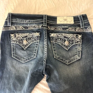 Miss Me jeans! - Snygga blå jeans från Miss Me med unika broderade bakfickor och dekorativa nitar. Jeansen är lågmidjade i en bootcut modell. Perfekta för dig som vill sticka ut med detaljer och cool stil. Nyskick! Midjemått: 38 cm, Innerbenslängd: 77cm, Storlek 27