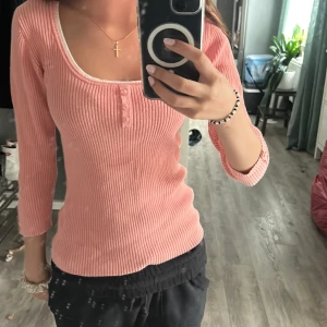 Rosa ribbad topp  - Superfin ljusrosa ribbad topp från papaya med trekvartsärm och knappar framtill. Djup rundad ringning med vit kant och smickrande slim fit. Perfekt att styla med jeans eller kjol för en chill look.