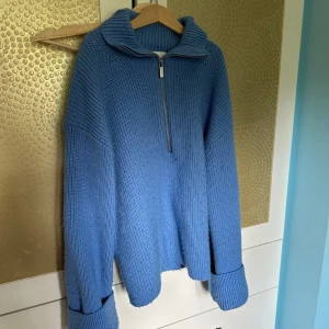 Blå stickad tröja med half zip - Mysig blå stickad tröja med hög krage och half zip-dragkedja framtill. Tröjan har en relaxed passform och långa ärmar med breda muddar. Perfekt för kyliga dagar när du vill vara både bekväm och snygg.