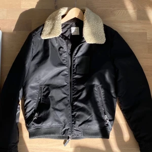 Svart bomberjacka med teddykrage från Ljung - Svart bomberjacka från Ljung i storlek M med schysst beige teddykrage. Storleken kan diskuteras då jag är mer av en S i passform. Jackan har dragkedja framtill, två snedställda fickor med dragkedja och ribbade muddar. Perfekt för dig som vill ha en stilren jacka med lite edge.