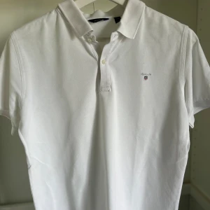 Vit pikétröja från GANT - Klassisk vit pikétröja från GANT med broderad logga på bröstet. Tröjan har krage, korta ärmar och knäppning med två knappar framtill. Tillverkad i bomull för en skön och luftig känsla. Perfekt för en clean och stilren look. Perfekt till dig som vill börja resella eller ha märkeskläder till feta priser