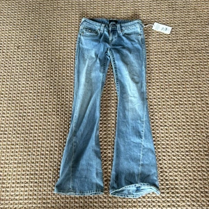 True Religion Joey Low Rise Flare jeans - Snygga ljusblå jeans från True Religion, modell Joey Low Rise Flare. De har låg midja, utsvängda ben och coola detaljerade bakfickor med silverknappar. Klassisk femficksdesign i mjuk bomullsdenim. Perfekta för dig som gillar retrovibbar och statement-jeans. De är helt sprillansnya och aldrig användt