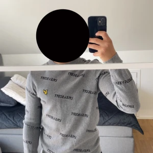 Grå stickad tröja Lyle & Scott M - Snygg grå stickad tröja från Lyle & Scott med svart logotyp-tryck över hela tröjan och den klassiska gula fågeln broderad på bröstet. Tröjan har rund halsringning, ribbade muddar och lång ärm. Perfekt för dig som gillar stilren streetwear.