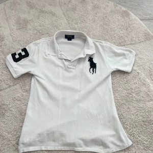 Vit pikétröja Polo Ralph Lauren  - Vit pikétröja från Polo Ralph Lauren med klassisk krage, svart broderad polospelare på bröstet och stor svart siffra 3 på ena ärmen.   har korta ärmar, perfekt för en clean och sportig look.