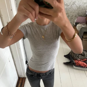 Grå ribbad t-shirt från H&M - En enkel och stilren grå ribbad t-shirt från H&M. Modellen är croppad med korta ärmar och rund halsringning. T-shirten är i mjuk bomull och har en tight passform som sitter snyggt på kroppen. Perfekt till jeans eller kjol för en clean look.