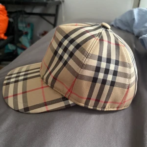 Burberry rutig keps i beige och svart - Säljer en klassisk Burberry keps i storlek L med ikoniskt rutigt mönster i beige, svart, vitt och röda linjer. Kepsen har justerbar snapback och svart insida. Märke med 'Burberry London England' baktill. Perfekt för dig som vill sticka ut med stil.