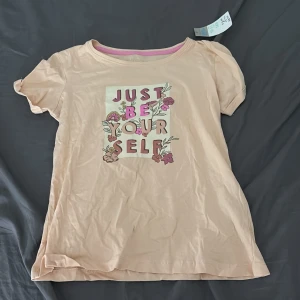 Beige t-shirt med blommor Primark - Söt beige t-shirt från Primark med tryck 'Just Be Yourself' och blommiga detaljer i rosa och lila. T-shirten har korta ärmar och är gjord i mjuk bomull. Perfekt för dig som gillar en chill och färgglad stil.