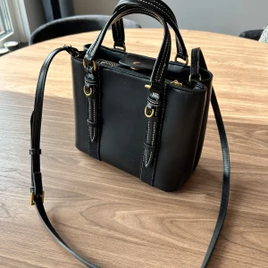 Svart väska med gulddetaljer  - Snygg svart axelväska i skinn med vita kontrastsömmar och guldfärgade metalldetaljer. Väskan har både handtag och lång axelrem, samt dragkedja och tryckknapp upptill. Perfekt storlek för det viktigaste och ger en stilren vibe.