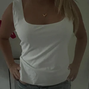 Vit ärmlös linnetopp med fyrkantig ringning - Säljer en stilren vit linnetopp med breda axelband och snygg fyrkantig ringning. Toppen är figurnära och passar perfekt till jeans eller kjol för en clean look. Enkel och fräsch design som är lätt att matcha.