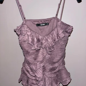 Ljuslila volangtop från Bik Bok - Supersöt ljuslila linnetopp från Bik Bok med tunna axelband och massor av volanger. Toppen har en rynkad struktur och volangdetaljer både fram och bak som ger en romantisk vibe. Perfekt för dig som gillar unika och lekfulla plagg.