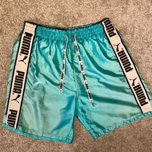 Turkosa shorts från Puma med logga - Snygga turkosa shorts från Puma med breda vita sidor där det står PUMA i svart. Shortsen har elastisk midja och snörning med Puma-tryck, samt meshfoder på insidan. Perfekta för sport eller chill dagar. Storlek XS eller junior 164