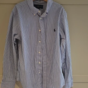 Blåvit randig skjorta Ralph Lauren - Snygg blå och vit randig skjorta från Ralph Lauren med klassisk krage och knappar framtill. Skjortan har lång ärm och ikonisk broderad logga på bröstet. Perfekt för dig som gillar stilrena och tidlösa plagg.