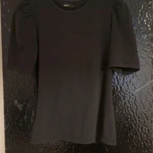 Svart topp med puffärm från ONLY - Svart topp från ONLY med puffade axlar och korta ärmar. Modellen är figurnära och har en klassisk rund halsringning. Perfekt att styla med både jeans och kjol för en trendig look.
