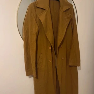 Lång brun kappa med knappar från Trendyol - Säljer en snygg lång kappa i brun färg med bred krage och dubbelknäppning framtill. Jackan har två fickor på sidorna och är perfekt för dig som gillar en stilren och klassisk look. Passar dig som vill ha en tidlös vibe i garderoben.