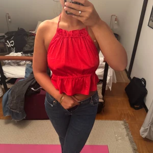 Röd halterneck topp med volang - Säljer en snygg röd halterneck topp med volangkant nertill och knyt i nacken. Toppen är ärmlös och har öppen rygg, perfekt för varma dagar. Materialet känns lätt och svalt, och den har en loose passform över magen.