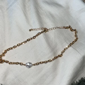Guldfärgat halsband med pärldetalj - Snyggt halsband i guldfärgad metall med grov kedja och en stor vit pärla som detalj. Justerbar längd med förlängningskedja och karbinlås. Perfekt accessoar för att lyfta din outfit.