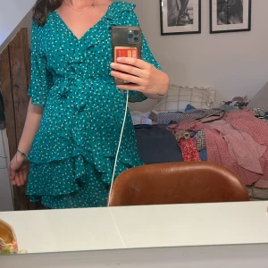 Turkos blommig omlottklänning - Superfin grön kortklänning med små vita och svarta blommor. Omlottmodell med volangdetaljer, v-ringning och knyt i midjan. Halvlånga ärmar och flera lager volanger ger en flowy känsla. Perfekt för sommarens alla tillfällen!