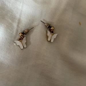 Söta fjärilsörhängen i vitt och guld - Ett par små örhängen med hängande fjärilar i vit och guldig metall. Framsidan har vit emalj och baksidan är helt guldig med struktur. Perfekta för dig som gillar gulliga detaljer och vill ha något unikt till din stil.