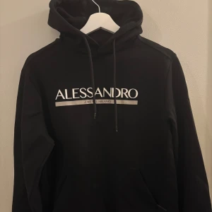 Svart hoodie från Alessandro - Svart hoodie från Alessandro Zavetti Milano med vit logga framtill. Klassisk modell med huva, snörning och känguruficka. Perfekt för en chill och stilren look. Mjuk bomullskänsla och långärmad design.