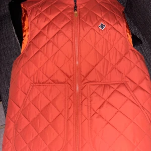 Quiltad orange väst från Morris - Snygg quiltad väst från Morris i en stark orange färg. Västen har två stora fickor framtill, dragkedja och en broderad logga på bröstet. Insidan är fodrad i glansigt orange tyg. Perfekt för lager-på-lager och ger en clean look. Helt ny väst.Priset kan alltid diskuteras.