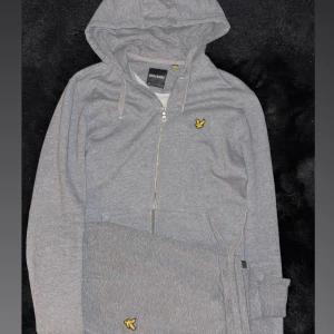Grå zip-hoodie dress från Lyle & Scott - Grå hoodie med dragkedja från Lyle & Scott, klassisk look med huva och snörning. Ikonisk gul logga på bröstet. Mjuk bomullskänsla, perfekt för chill dagar eller när du vill vara bekväm men ändå snygg. Fickor framtill och ribbade muddar.