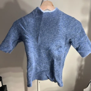 Blå stickad tröja från Holzweiler - Supermjuk blå stickad tröja från Holzweiler med korta ärmar och hög hals. Tröjan har en fluffig känsla och är perfekt för lager-på-lager. Enkel och stilren design som passar till många olika looks.