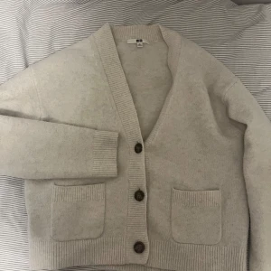 Beige kofta från Uniqlo XS - Mysig beige kofta från Uniqlo i storlek XS. Koftan har djup v-ringning, stora bruna knappar och två praktiska fickor framtill. Ribbstickade muddar och kant ger en clean look. Perfekt för lager-på-lager under höst och vinter.